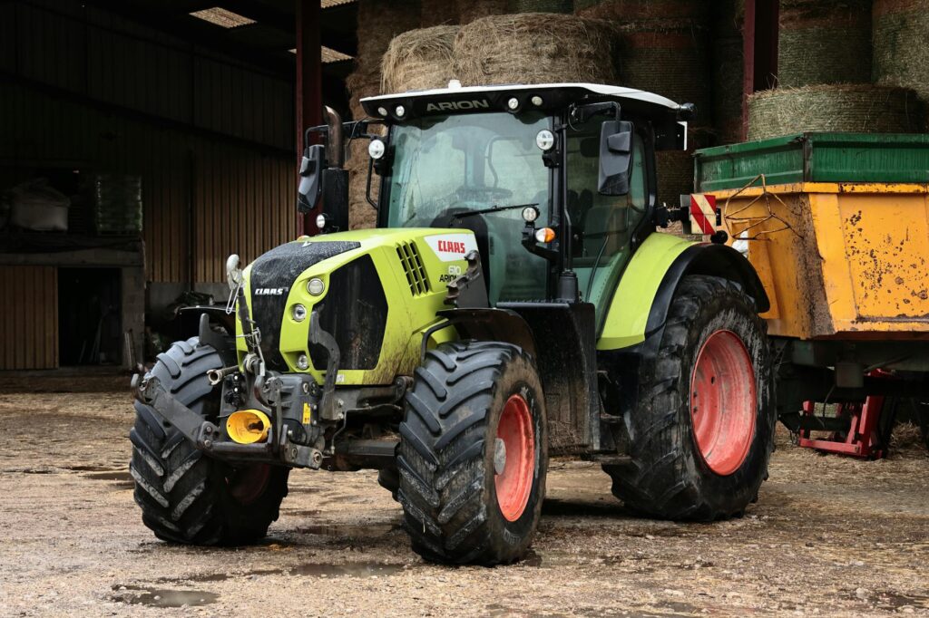 A green and yellow tractor parked in a farm with a trailer and bales of hay.